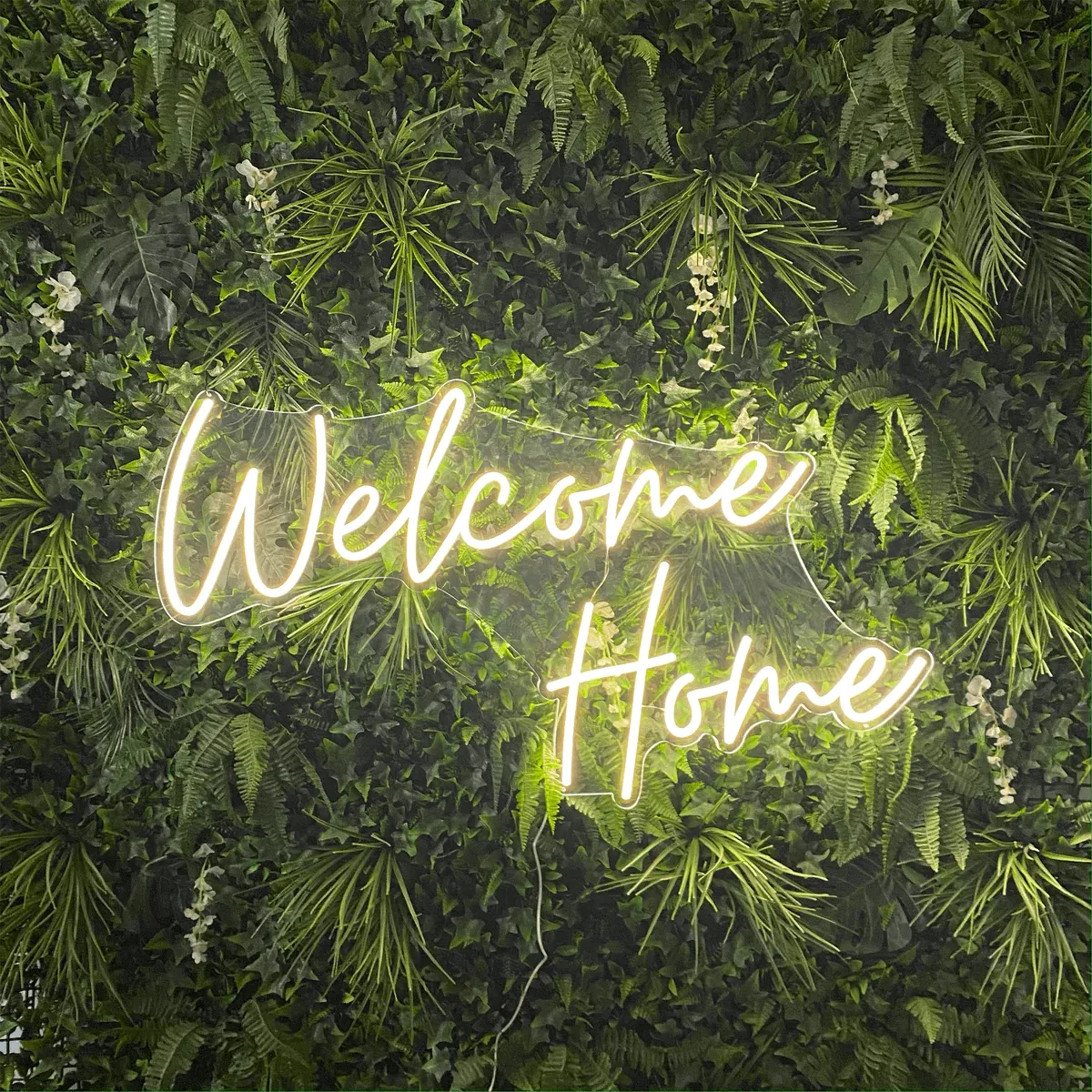 A warm-white 'Welcome Home' neon script sign mounted on a green plant wall
