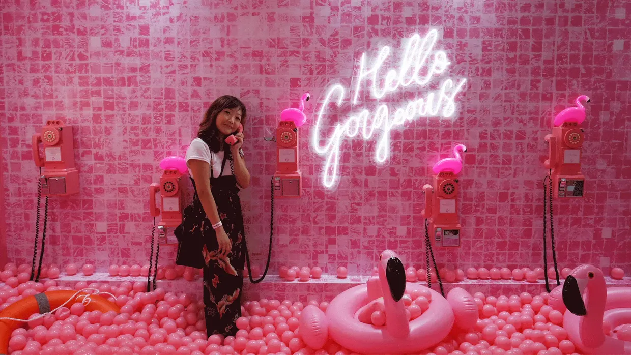 A person standing in a pink ball pit beside a white cursive neon sign reading Hello Gorgeous